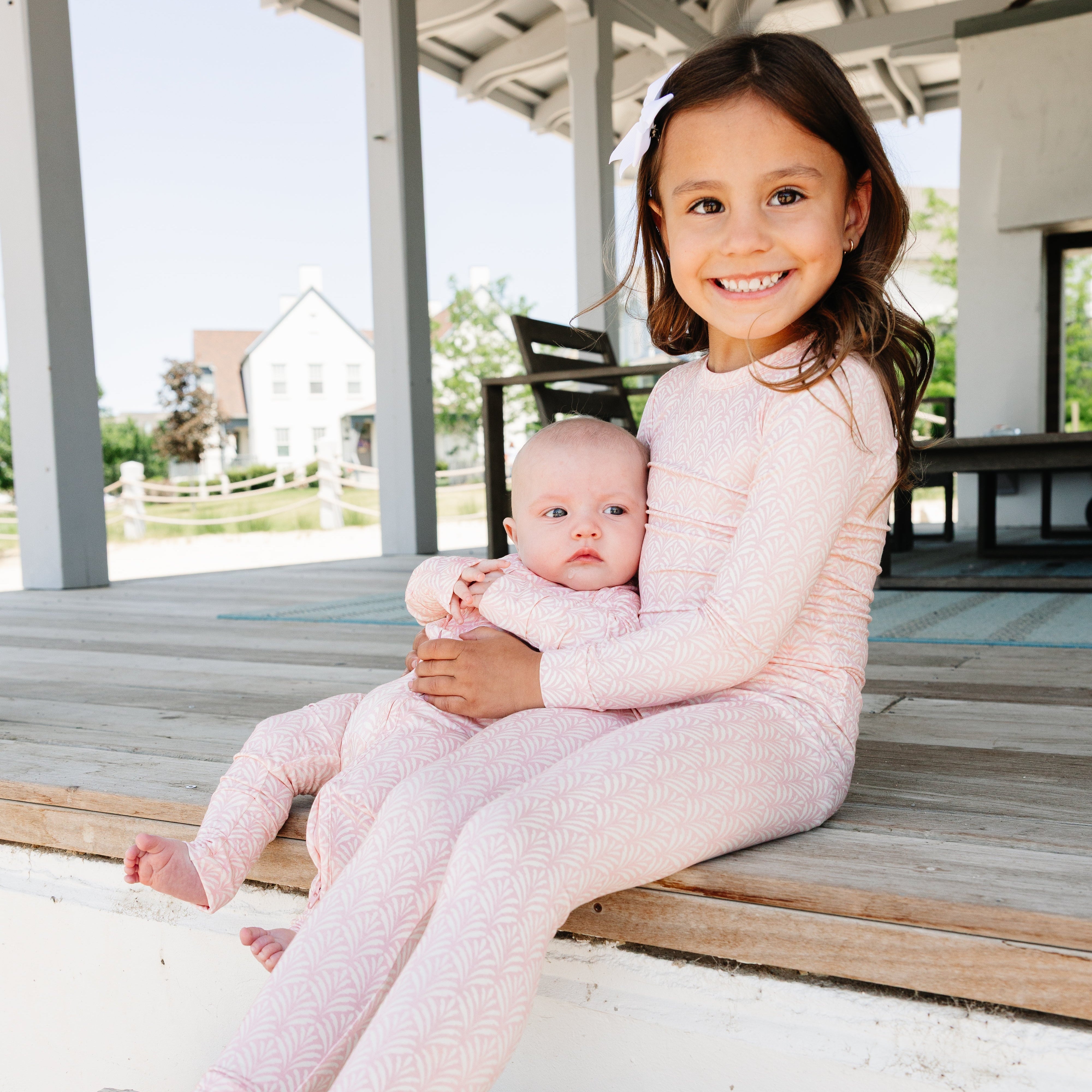 Two children in matching pink bamboo pajamas sitting on a wooden deck.