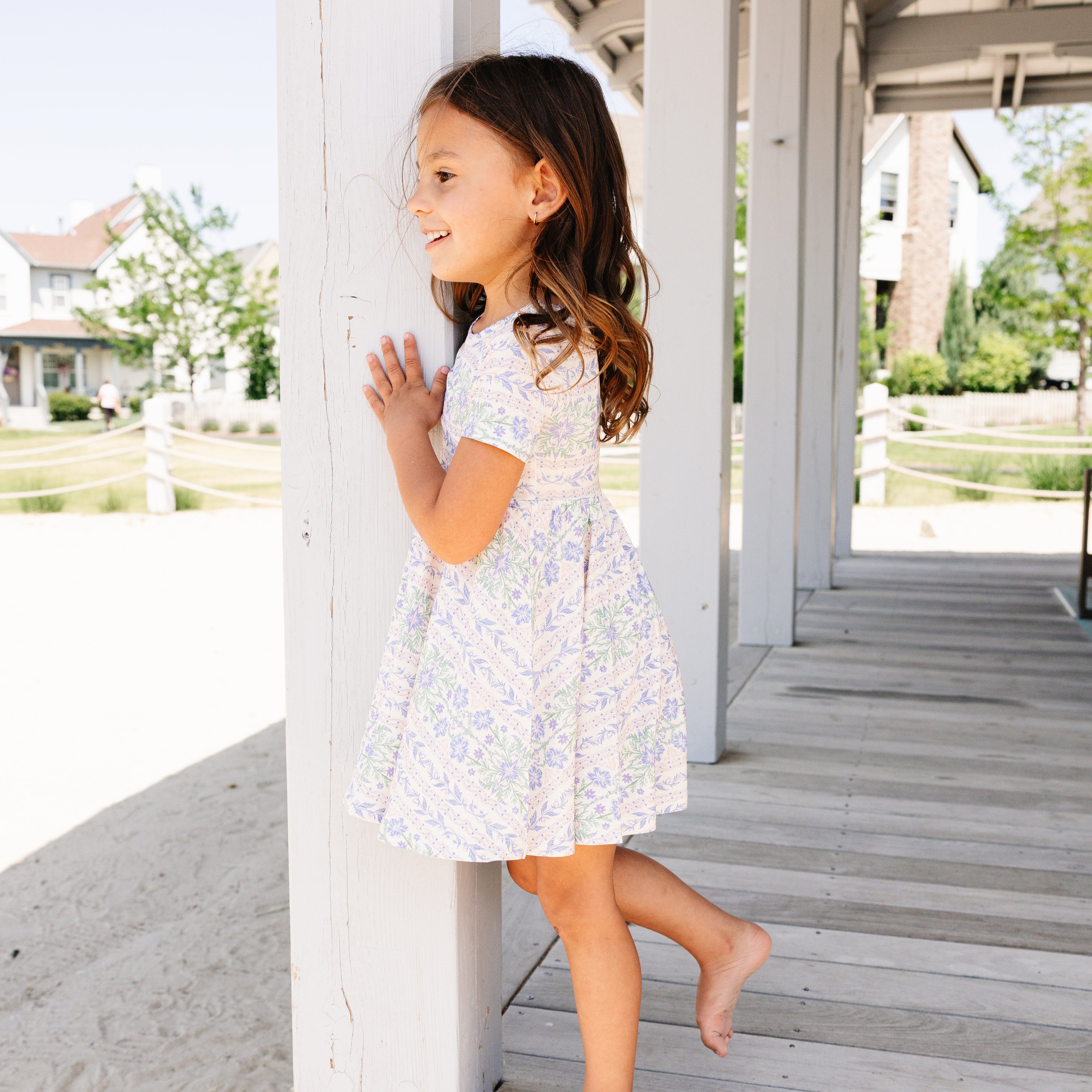 Young girl in a floral bamboo twirl dress standing on a wooden deck with a beach and house in the background.
