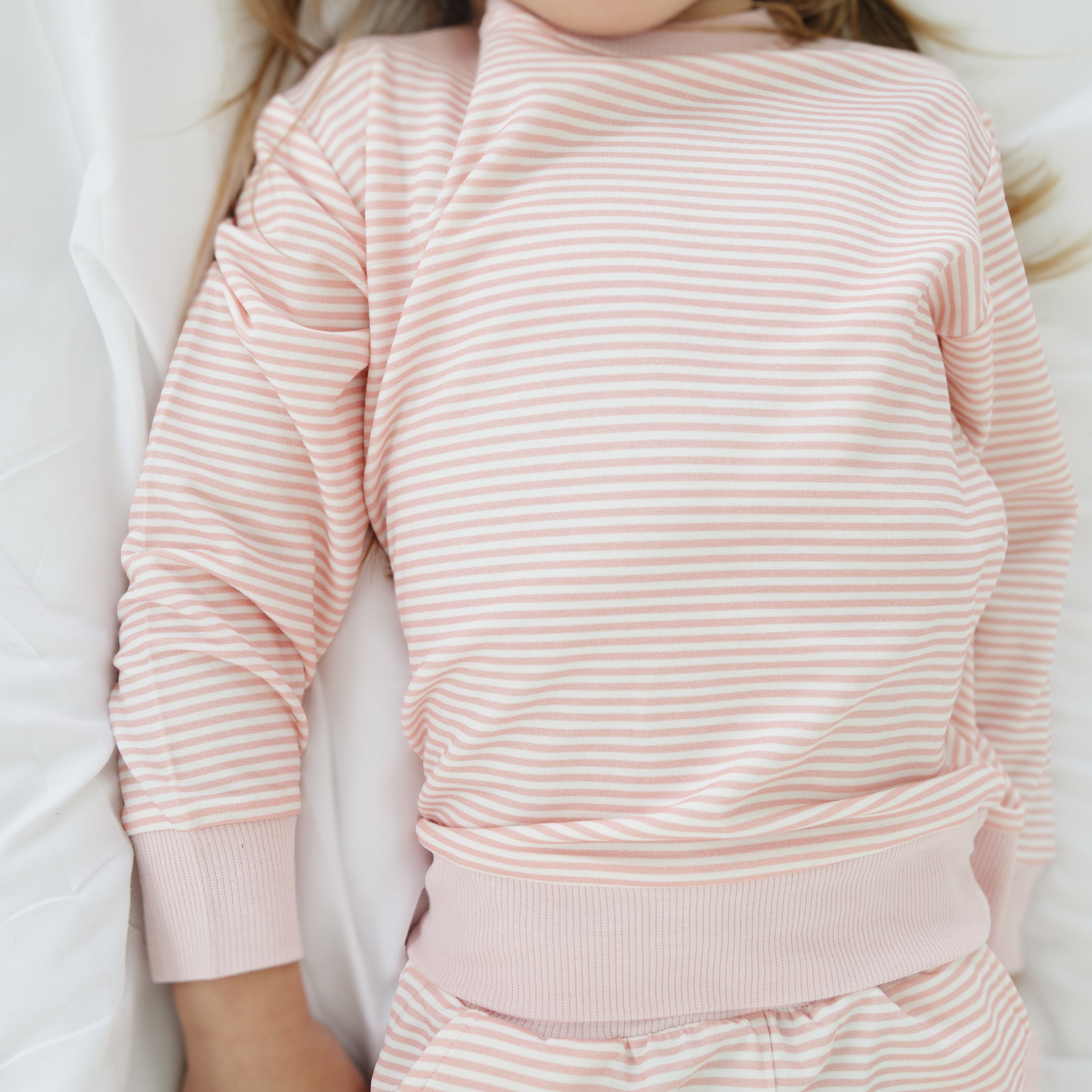 Girl laying in bed wearing the pink striped bamboo sweatsuit, resting peacefully under a light blanket