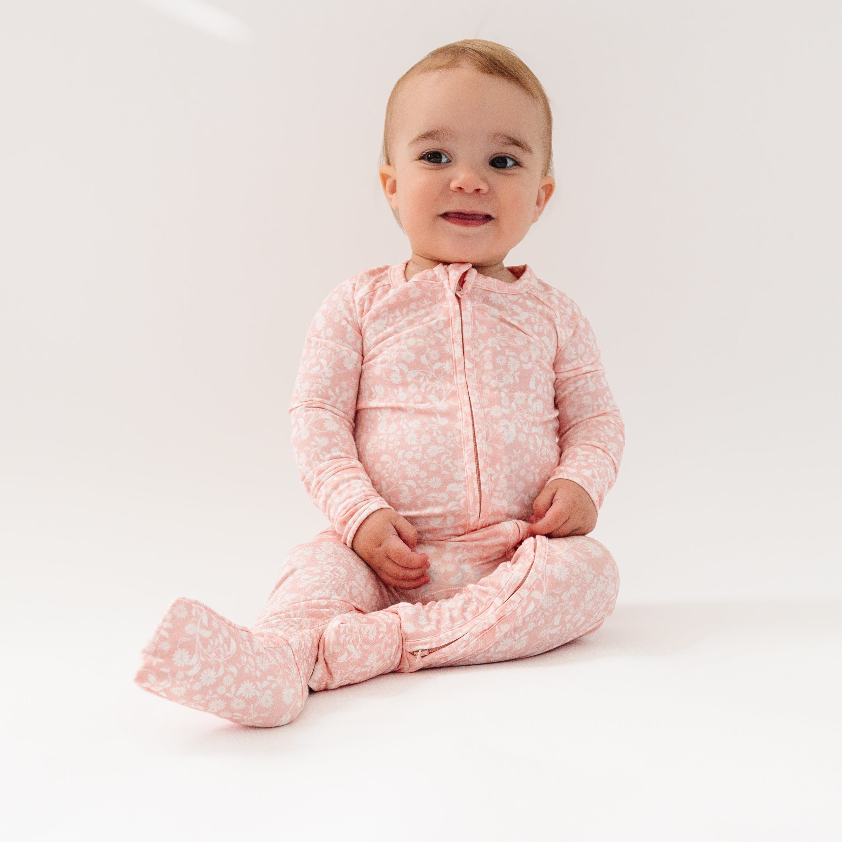 Little girl sitting down in Motette’s pink floral bamboo sleeper, smiling softly with hands in her lap
