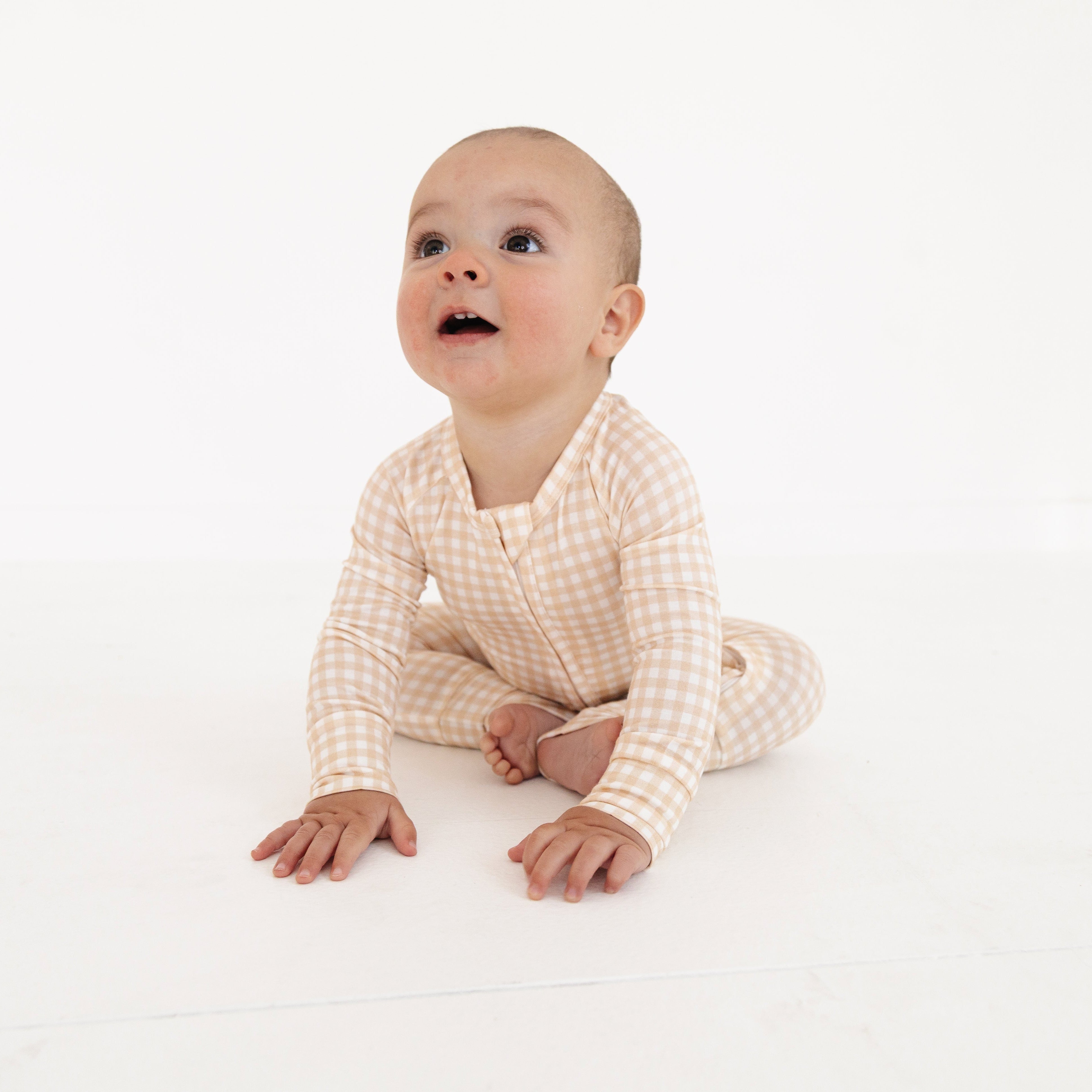 Newborn baby wearing beige checkered bamboo zipper romper, sitting upright and looking curious.