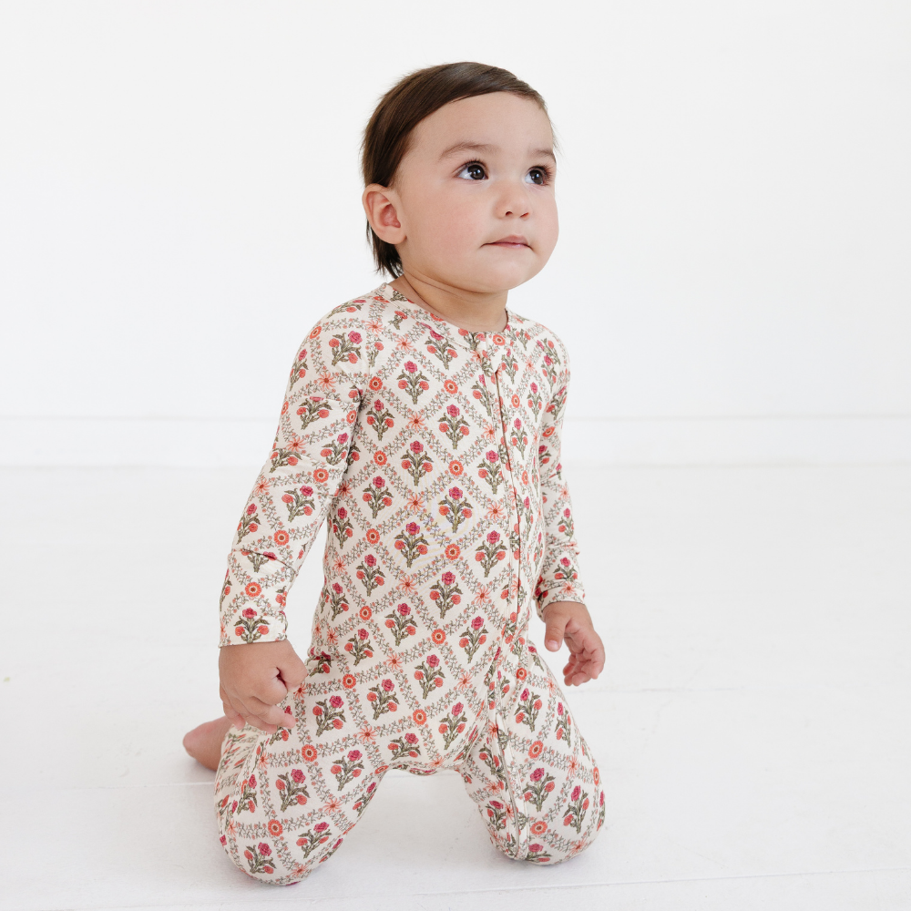 Baby kneeling on a white floor and looking upward wearing the Motette Holly Trellis Bamboo Sleeper, highlighting the comfortable fit and intricate vintage floral pattern.