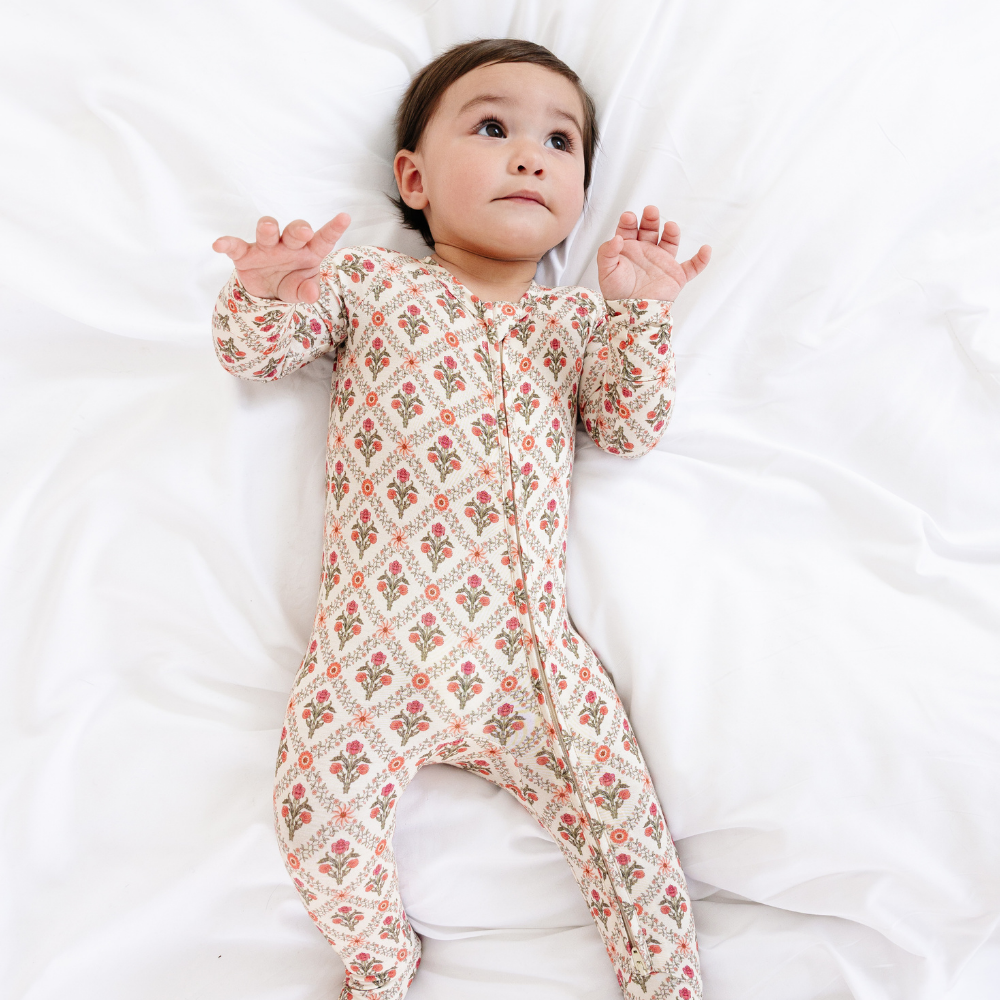Baby lying on a white surface with arms raised wearing the Motette Holly Trellis Bamboo Sleeper, showing the full-body coverage and vibrant coral floral trellis print.