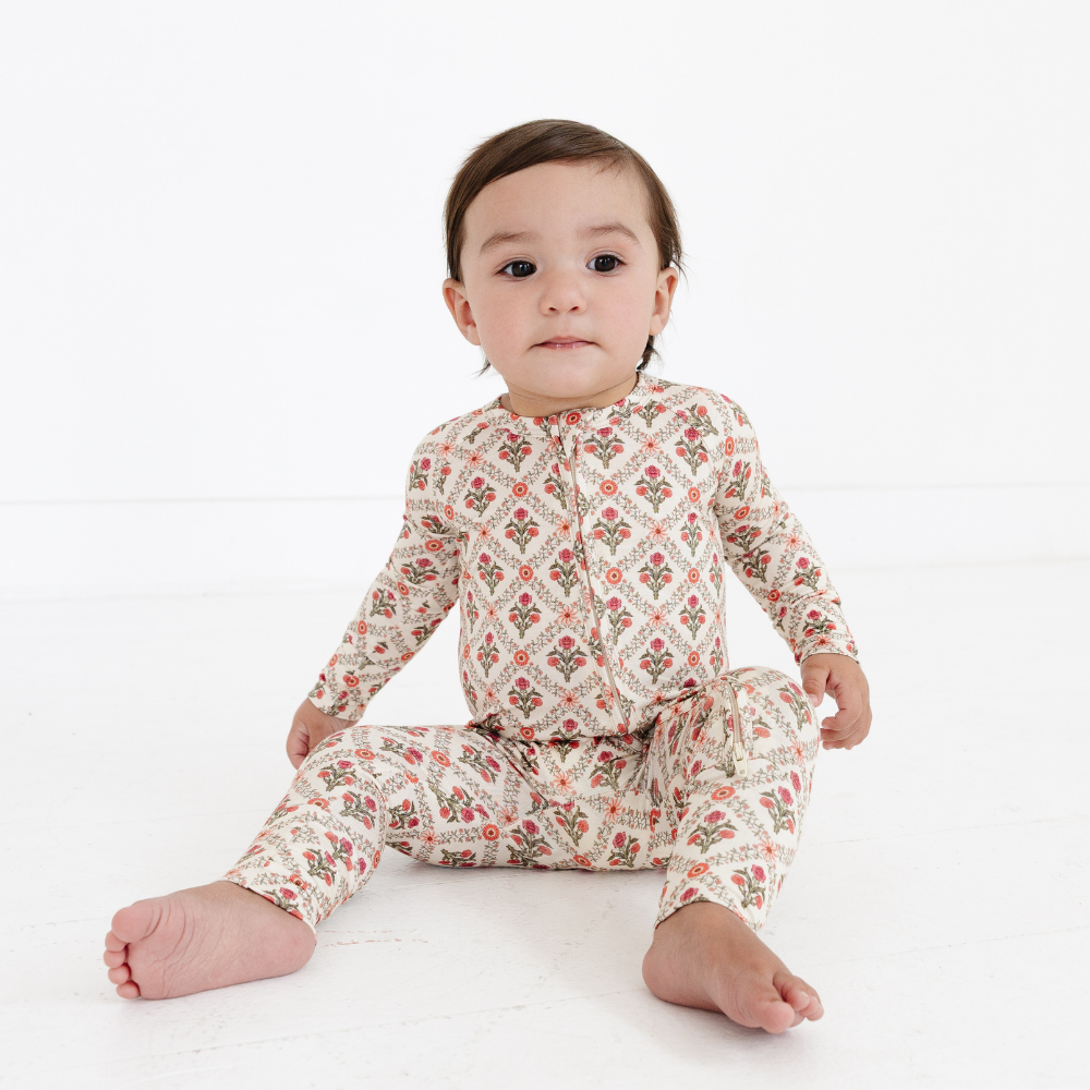 Baby sitting on a white floor wearing the Motette Holly Trellis Bamboo Sleeper, featuring a cream base with a coral floral trellis pattern, long sleeves, and a functional double zipper.