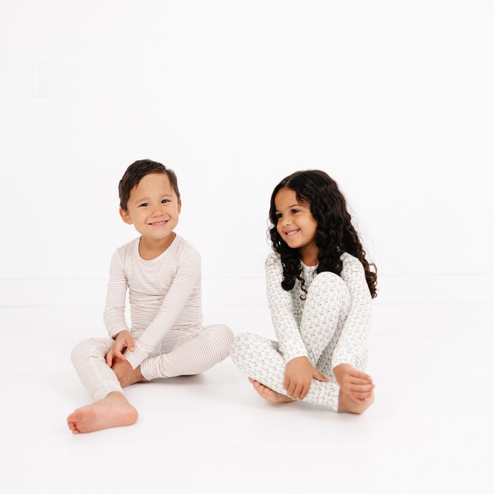 Two kids sitting on floor smiling and laughing, one wearing Motette Lavender Lane Bamboo Pajama Set, the other in Oatmeal Stripe.