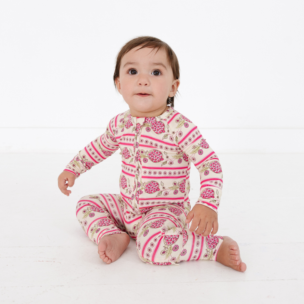 Baby sitting and looking to the side wearing the Motette Pomegranate Soiree Bamboo Sleeper, featuring a cream base with pink pomegranates, florals, and stripes, a double zipper, and fitted cuffs.