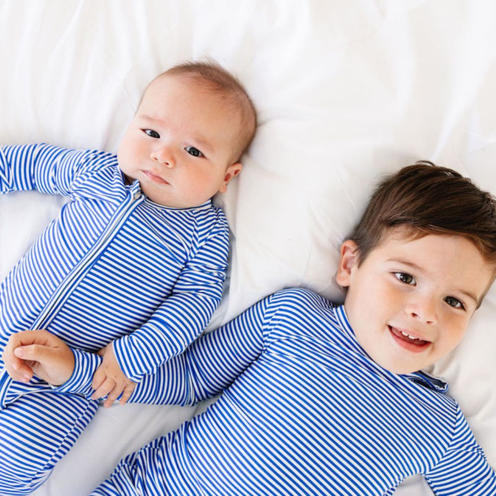 Toddler and baby lying on a white bed in matching Cobalt Stripe bamboo pajamas by Motette – breathable and eczema friendly sibling sleepwear