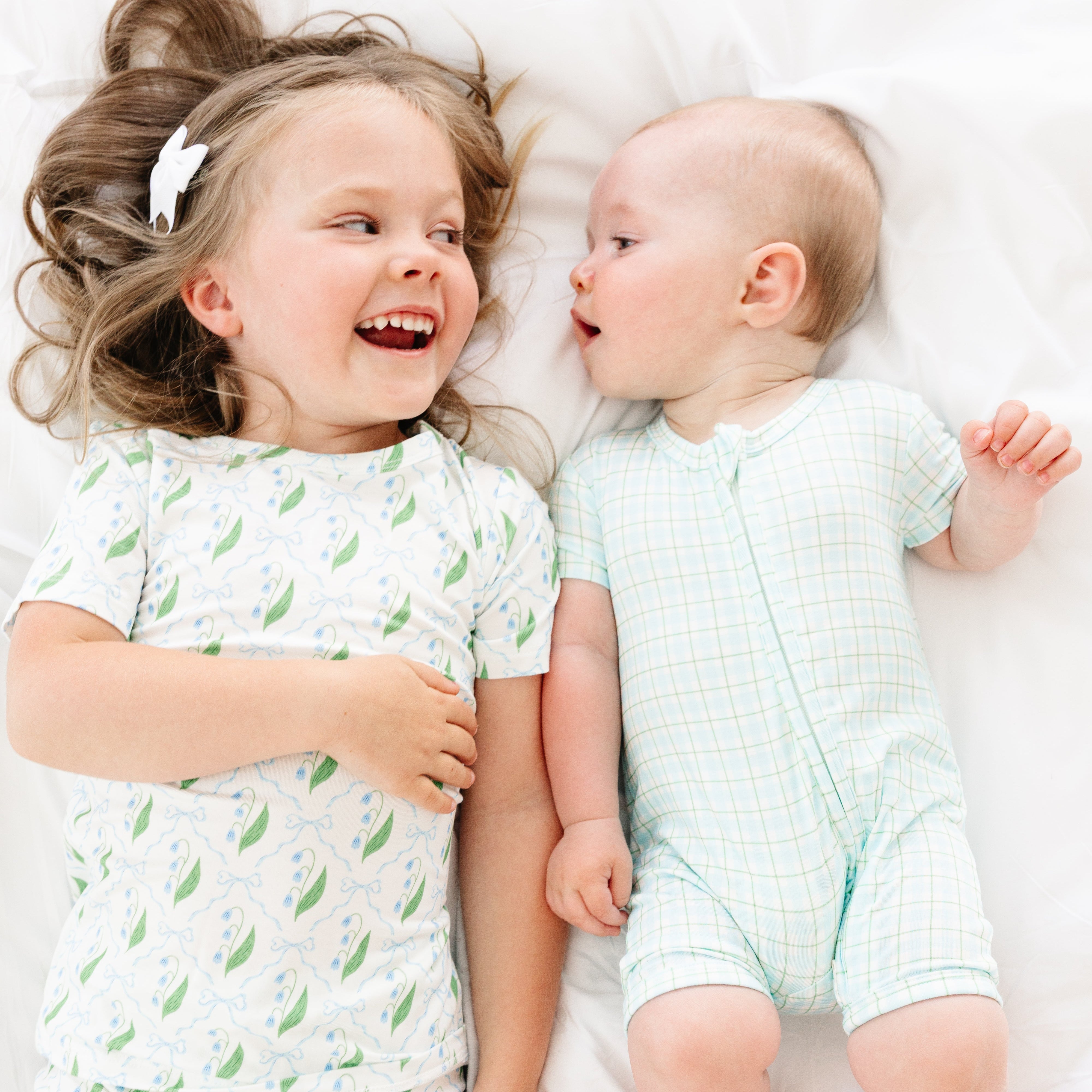 Toddler girl and baby sister lying down and smiling at each other in matching Bluebell Bows bamboo pajama sets from Motette