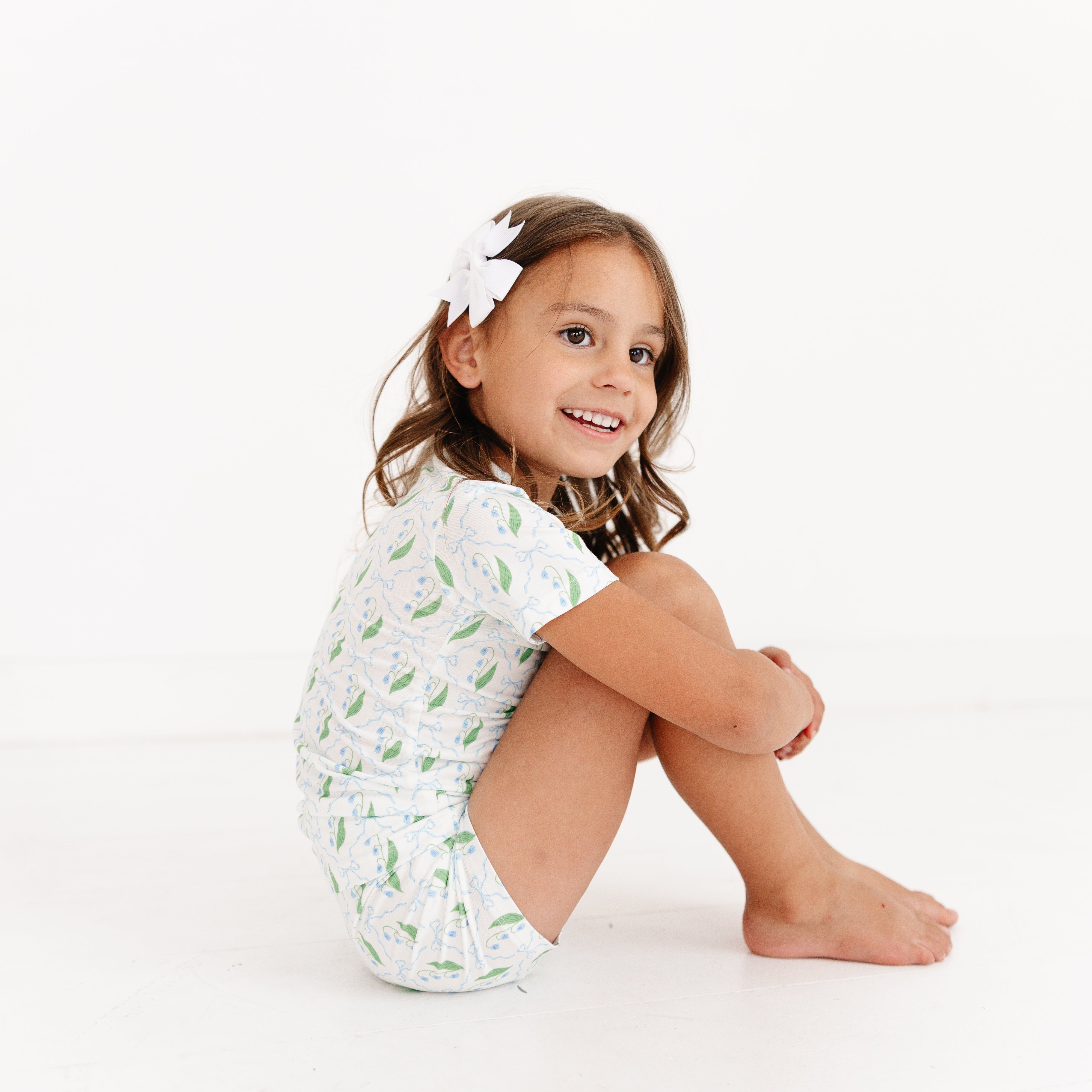Toddler girl smiling while sitting and wearing the Bluebell Bows Short Sleeve & Shorts Bamboo Toddler Pajama Set by Motette