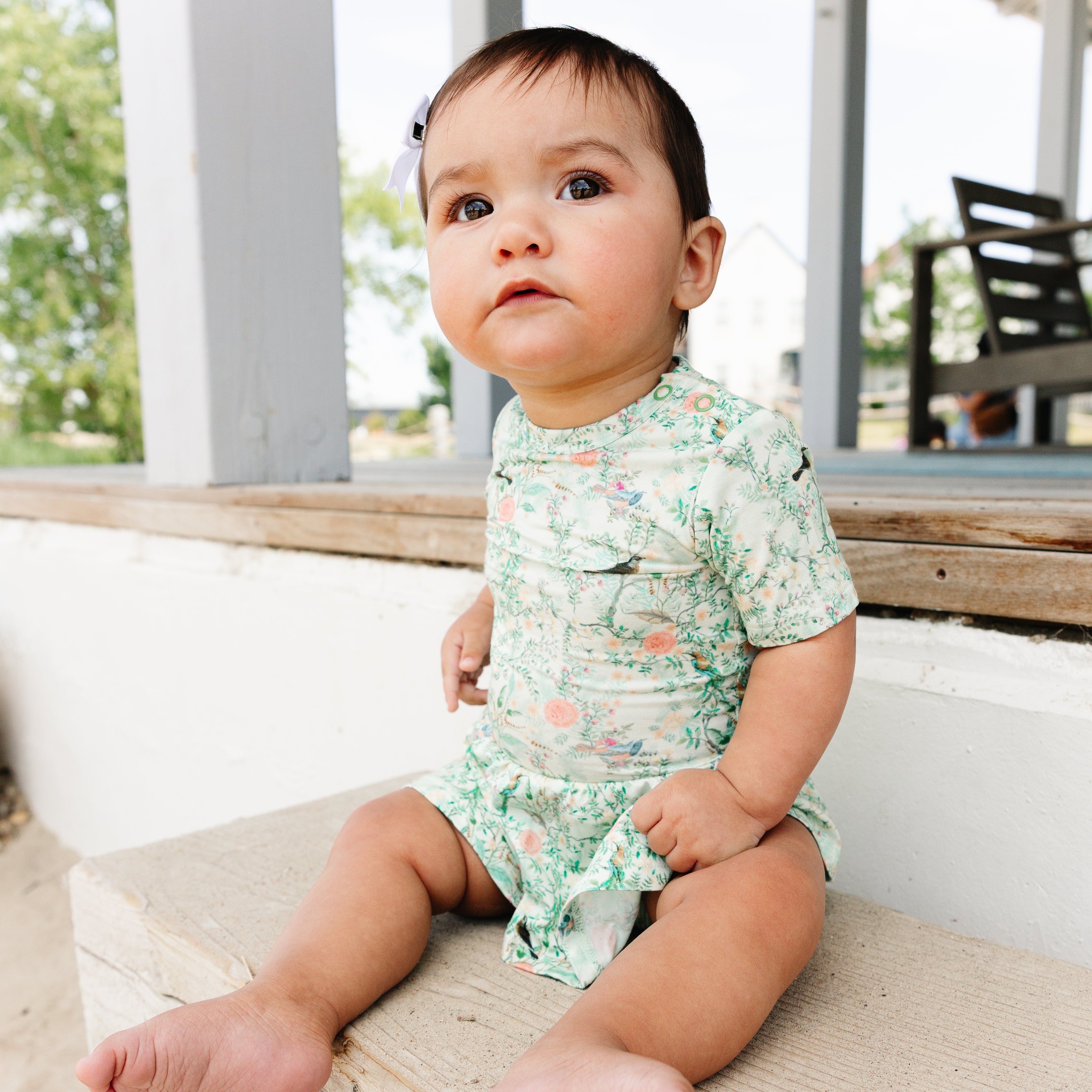 Baby wearing a green patterned top ranked bamboo twirl dress sitting on a wooden surface outdoors.