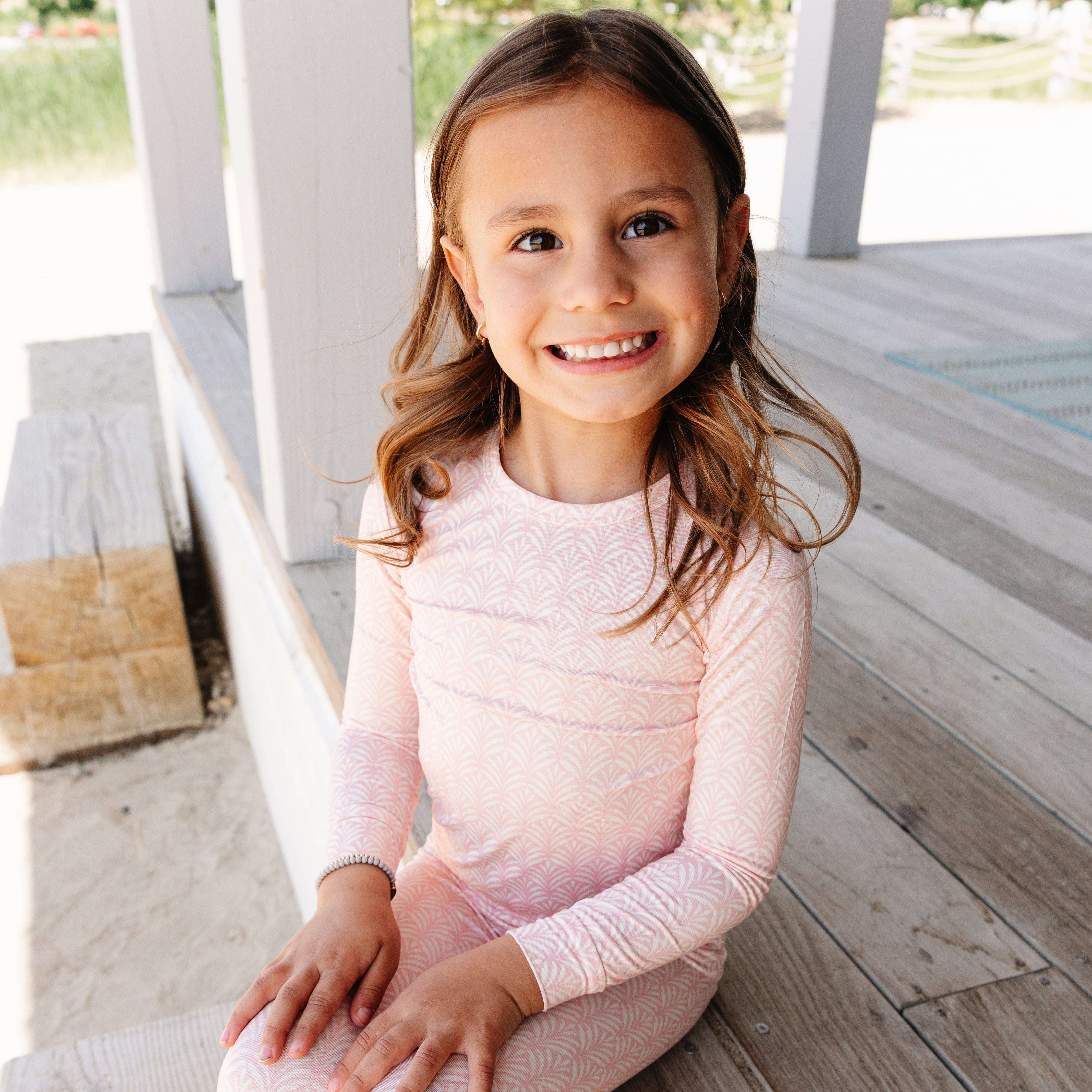 Young girl in top ranked pink bamboo pajamas sitting on a wooden deck.