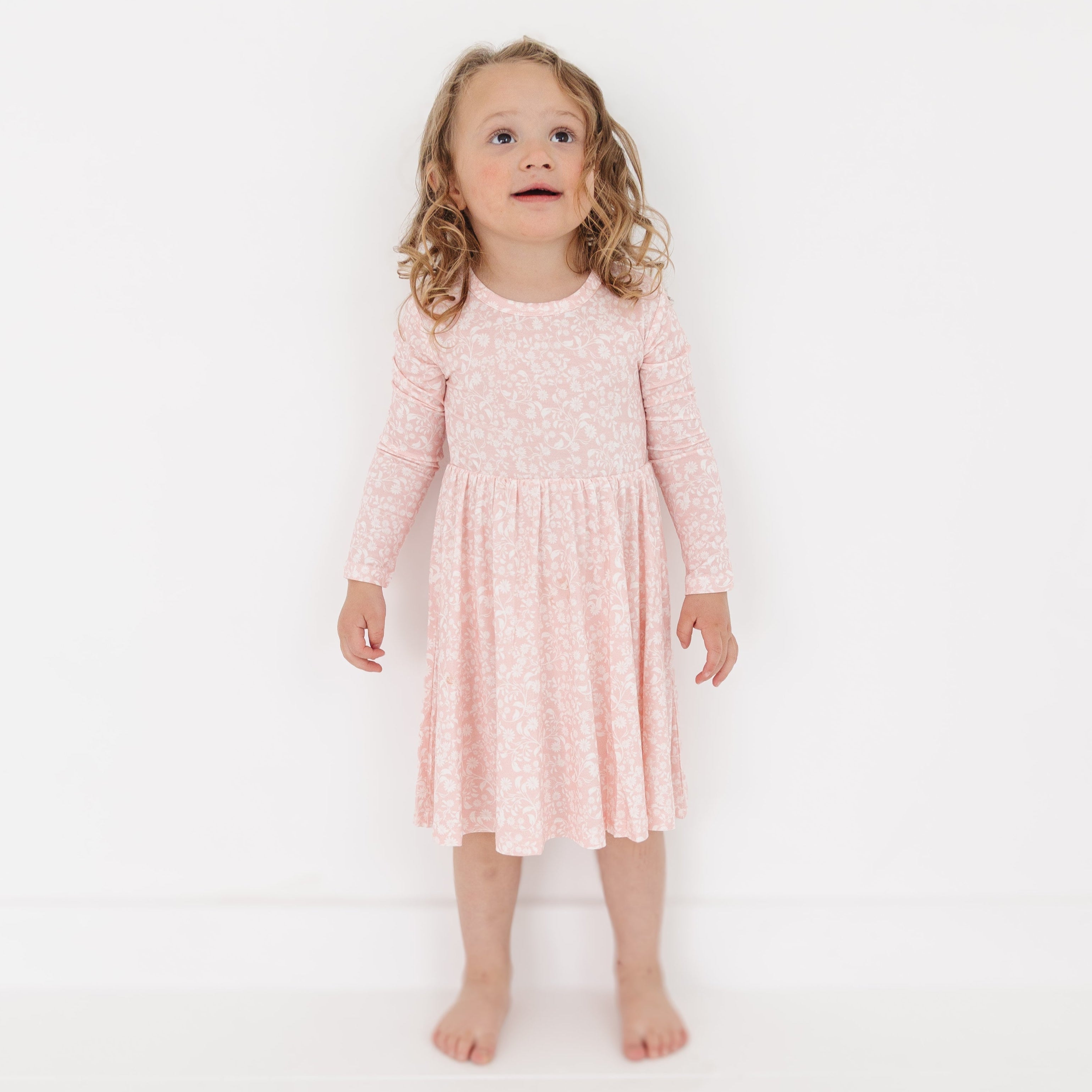 Girl standing in Motette’s long-sleeve pink floral bamboo twirl dress, smiling with her hands by her sides