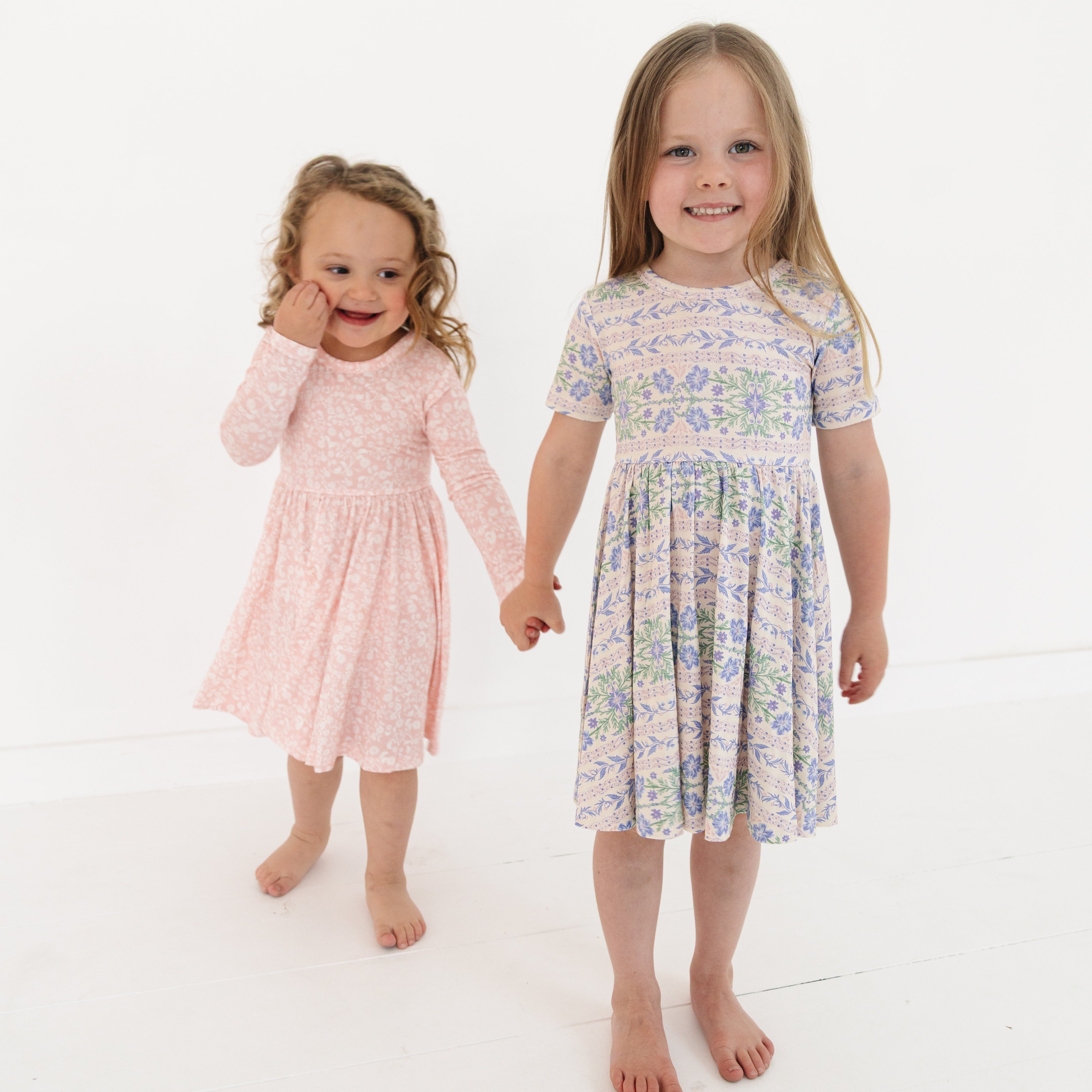 Sisters wearing coordinating pink floral bamboo twirl dresses, holding hands and smiling at each other