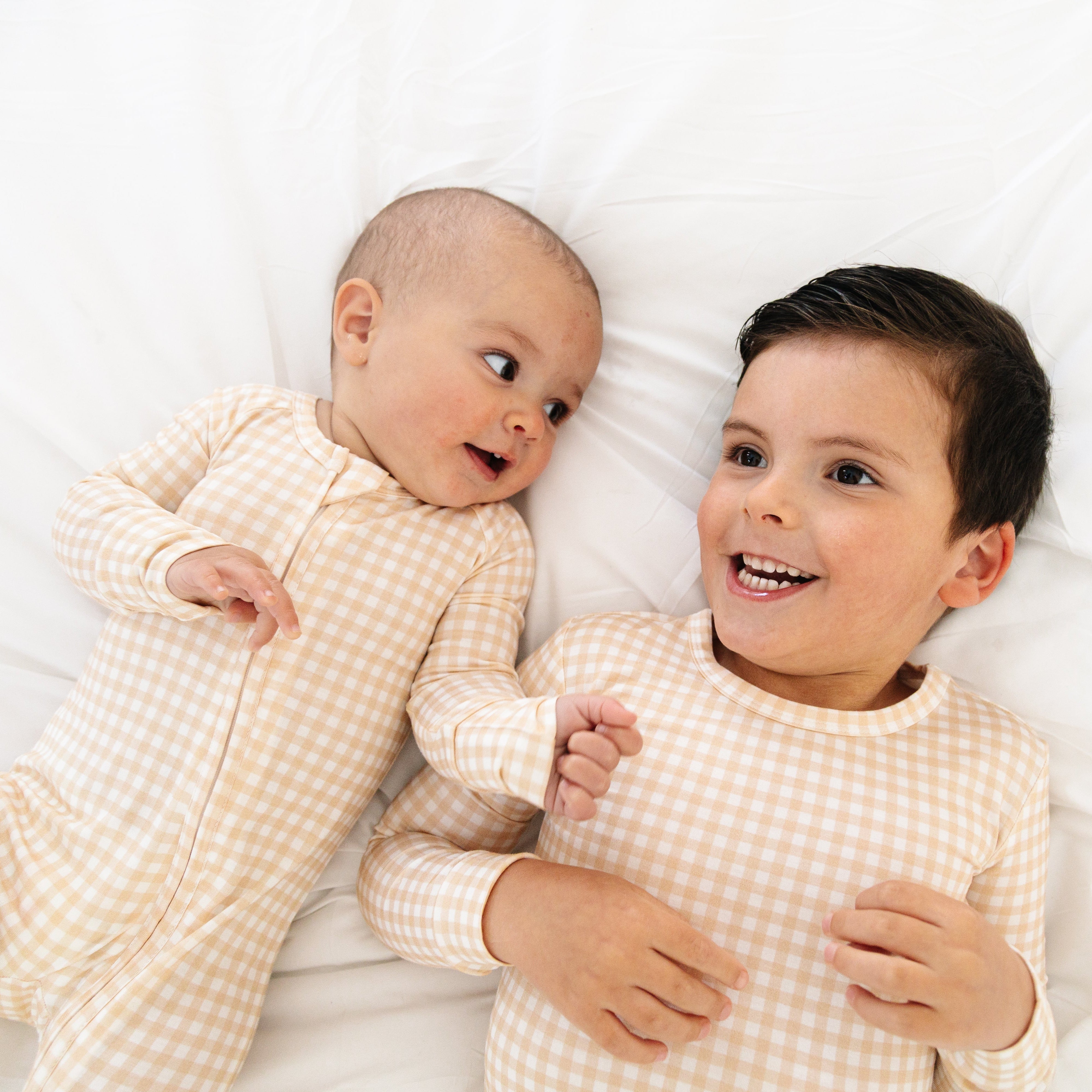 Close-up of newborn and toddler in beige checkered bamboo sleeper and two piece pajamas laughing and smiling