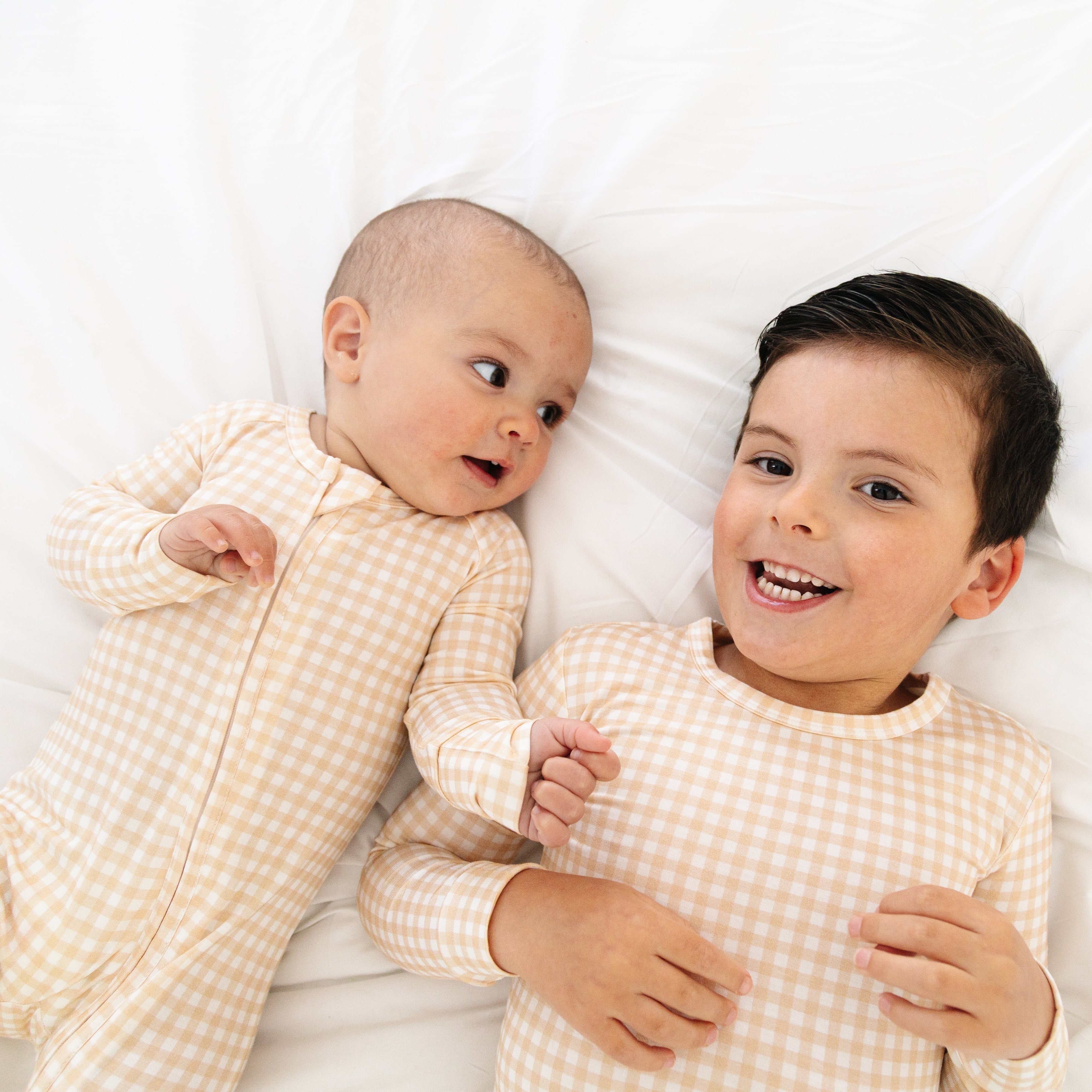Toddler boy and baby brother wearing matching beige checkered bamboo pajamas—two-piece and sleeper styles—laying together
