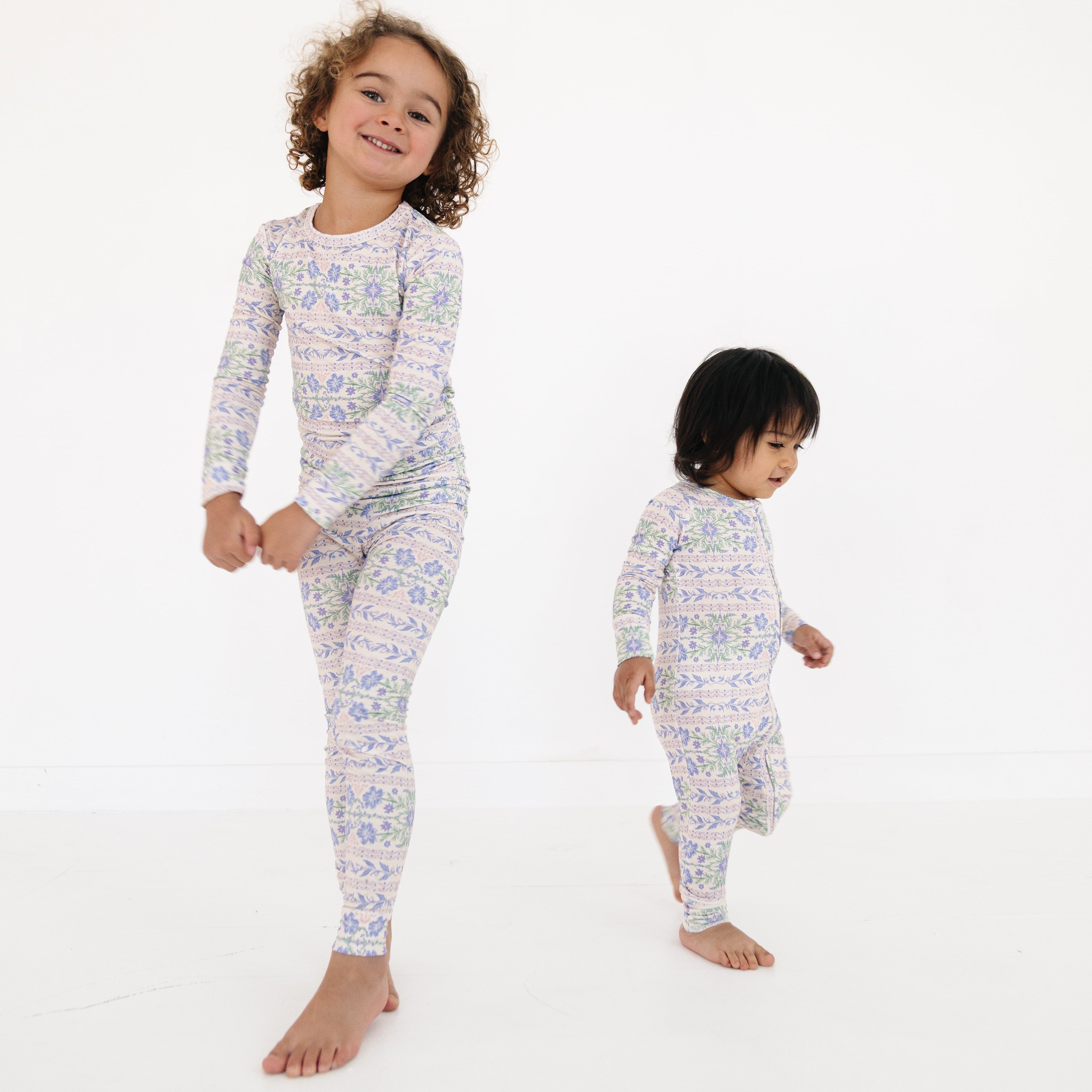 Siblings standing together wearing coordinating Motette bamboo pajamas—toddler in the two-piece set and baby in the matching sleeper