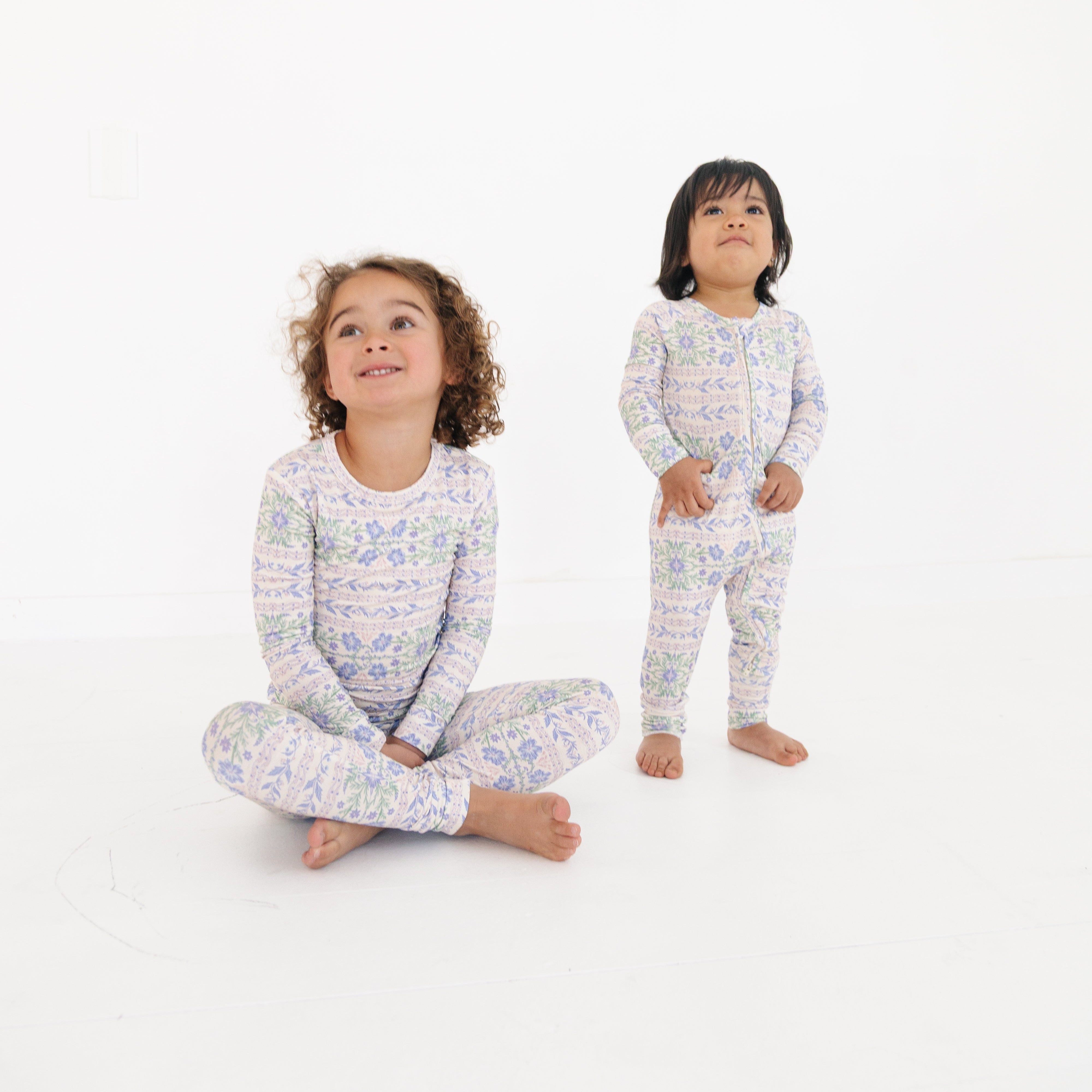 Siblings in matching pink and purple floral bamboo PJs—baby in sleeper, toddler sitting cross-legged in the two-piece set