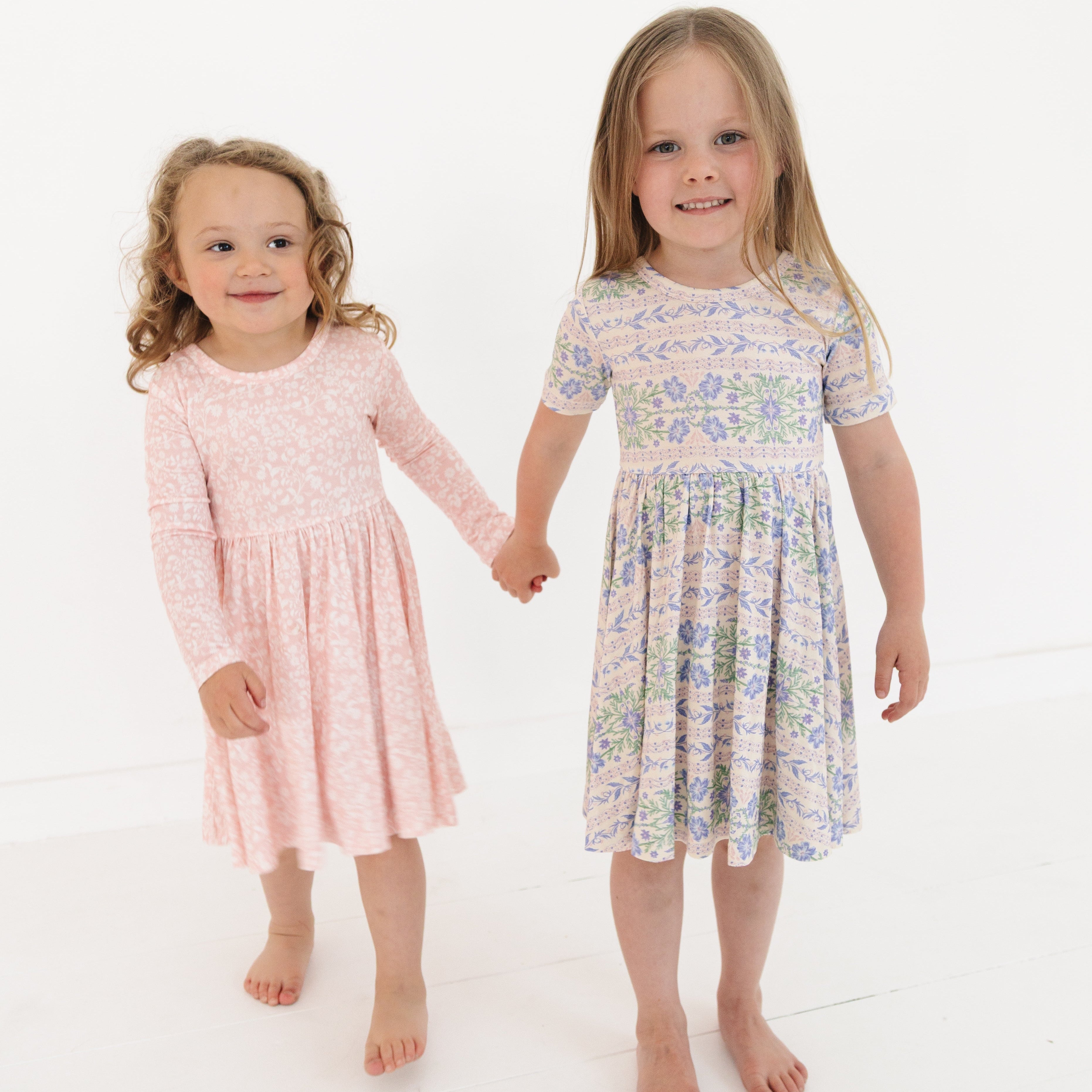 Sisters in matching pink and purple floral bamboo twirl dresses, standing side by side and holding hands