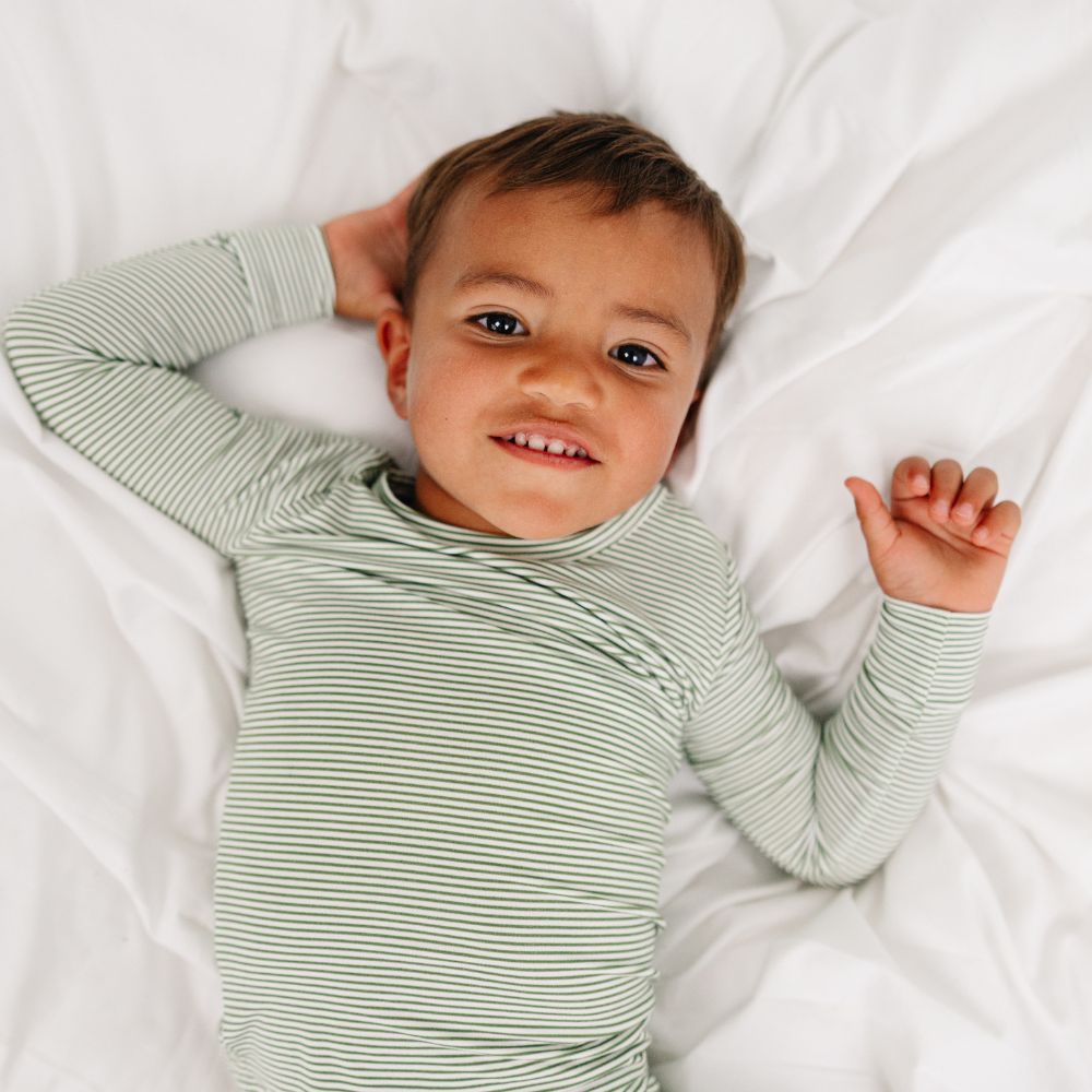 Child lying on bed wearing Motette Evergreen Stripe Bamboo Kids Pajama Set with green stripe pattern.
