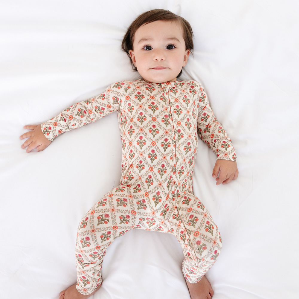 Baby lying on their back on a white sheet wearing the Motette Holly Trellis Bamboo Sleeper, showcasing the soft bamboo fabric and the repeating red and green floral trellis design.