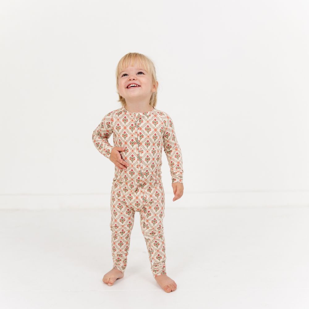 Baby standing and smiling wearing Motette Holly Trellis Bamboo Sleeper with red floral pattern.
