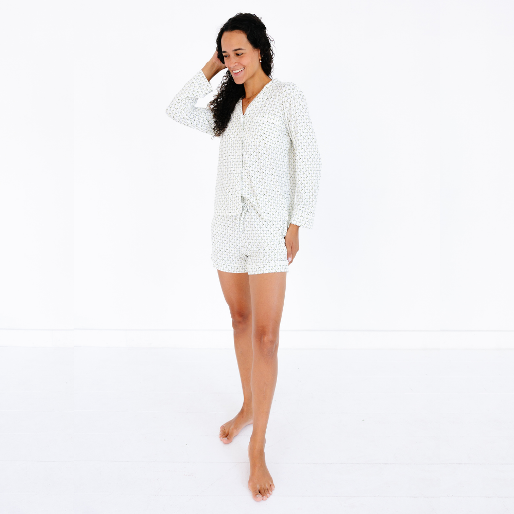 Mommy standing with one hand in her hair and looking down wearing the Motette Lavender Lane Bamboo Women's Pajama Set, featuring a long-sleeve button-up top and matching shorts with a soft cream base and delicate lavender floral pattern.