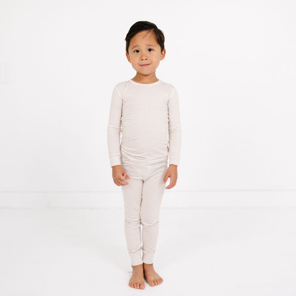 Young boy standing front-facing wearing Motette Oatmeal Stripe Bamboo Kids Pajama Set, smiling softly.