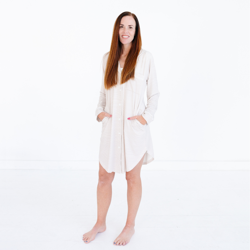 Woman standing and smiling wearing the Motette Oatmeal Stripe Bamboo Women's Nightgown, featuring thin tan and cream horizontal stripes, a full button-down front, long sleeves, and a relaxed mid-thigh fit with hands tucked into side pockets.
