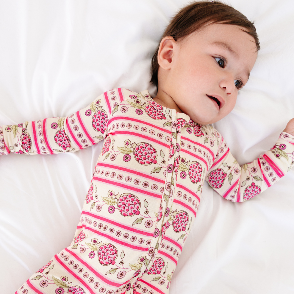 Close-up of a baby lying on their back wearing the Motette Pomegranate Soiree Bamboo Sleeper, highlighting the soft bamboo fabric, detailed pink pomegranate print, and double zipper closure.