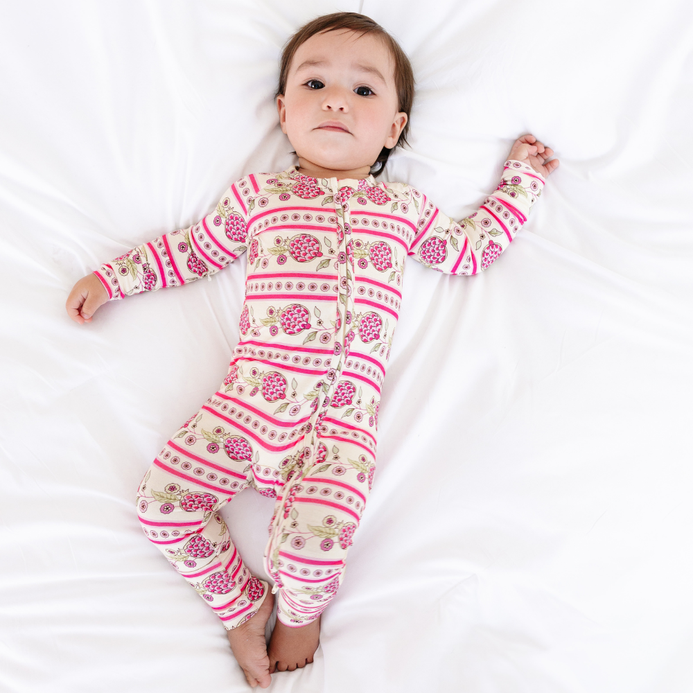 Full body view of a baby lying on a white sheet wearing the Motette Pomegranate Soiree Bamboo Sleeper, featuring the vibrant pink pomegranate print, double zipper, and convertible foot cuffs.
