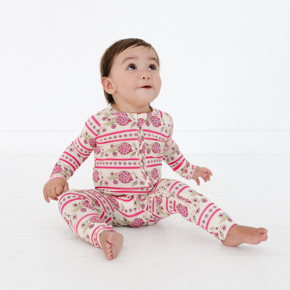 Baby sitting and looking up wearing the Motette Pomegranate Soiree Bamboo Sleeper, showing the full pink pomegranate and stripe pattern, snug fit, and long sleeves.