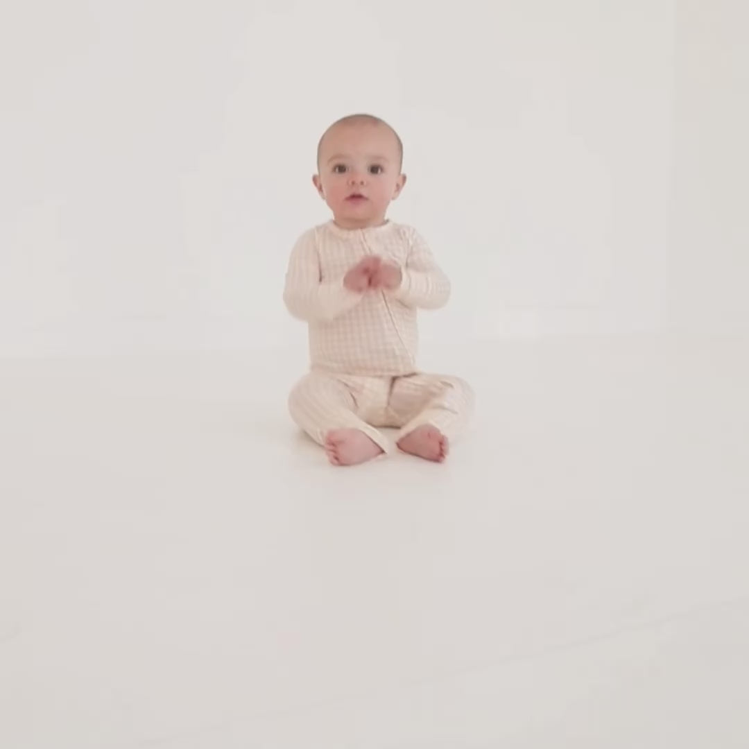 Baby and sibling video wearing beige checkered bamboo zipper romper, sitting upright and looking curious.