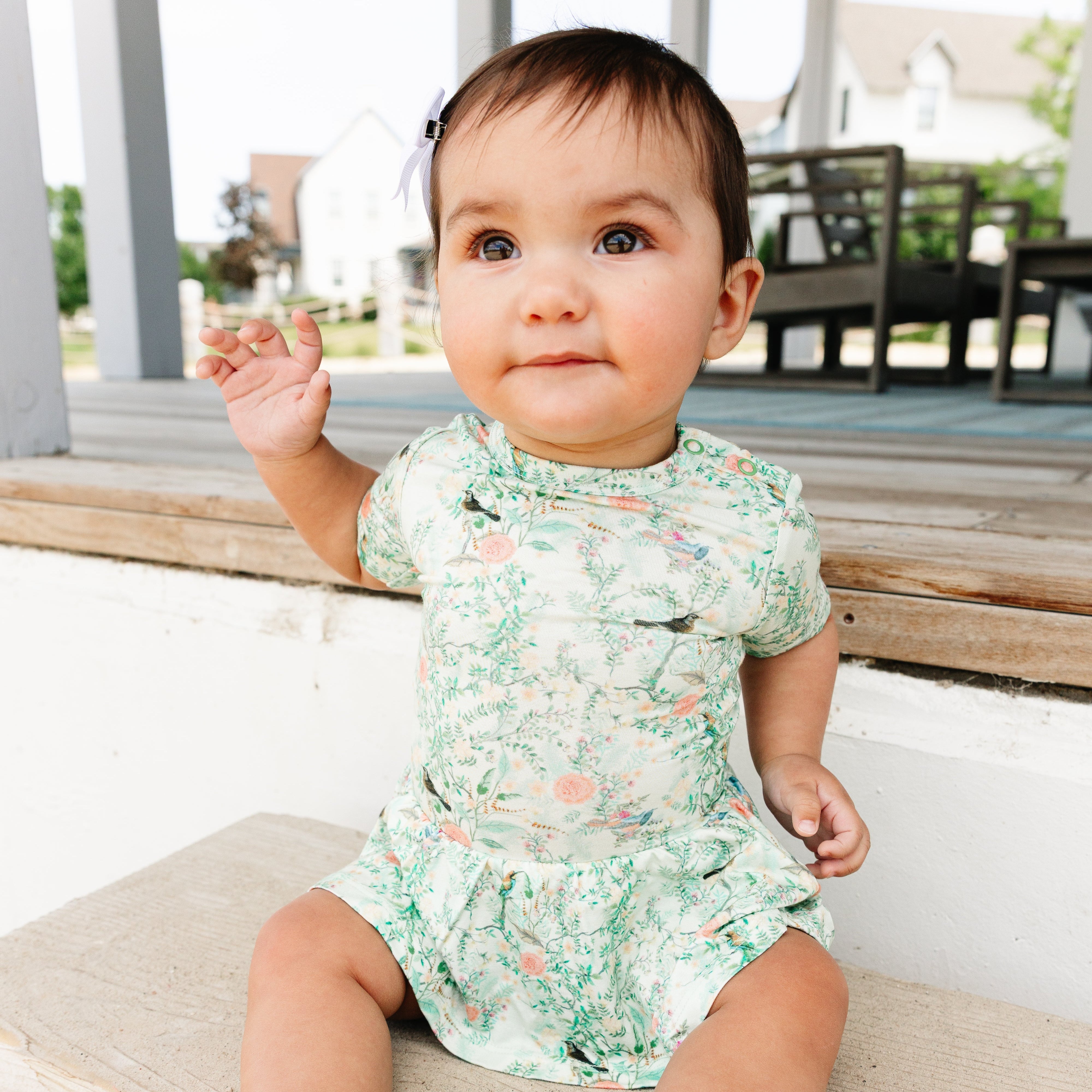 Baby in a floral twirl dress sitting on a wooden deck.